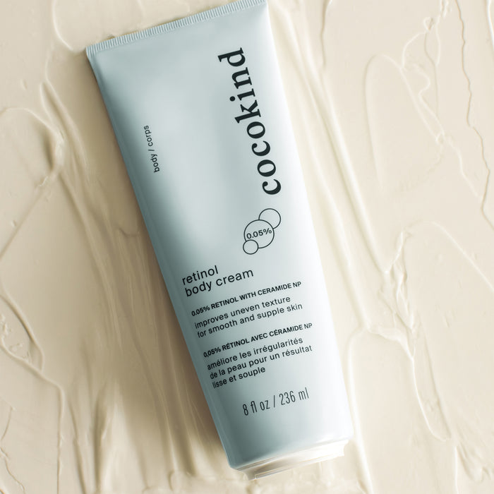 An 8 fl oz (236 ml) Cocokind Retinol Body Cream in a light blue tube sits on a textured surface. The label highlights squalane, 0.05% retinol, and ceramide NP as key ingredients for skin nourishment.