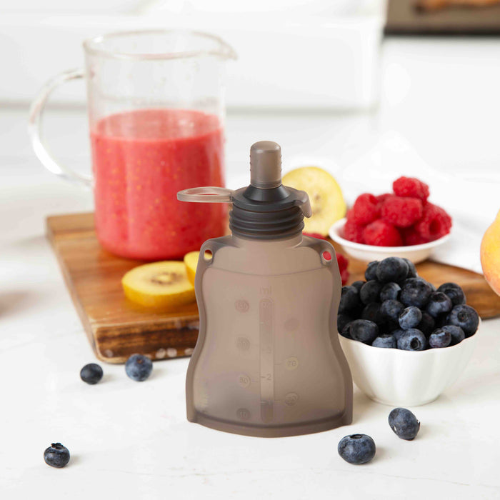 The Haakaa Reusable 100% Food Grade Silicone Baby Food Pouch sits on the kitchen counter among vibrant fruits. Nearby, a clear container is filled with red smoothie, and a white bowl holds blueberries next to a wooden board adorned with raspberries and sliced kiwi for your self-feeding journey.