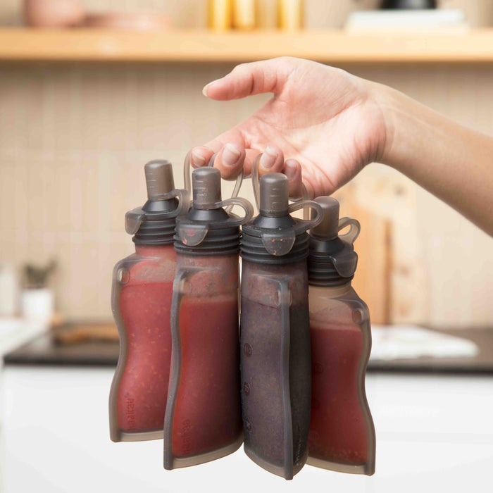 A person holds four Haakaa Reusable 100% Food Grade Silicone Baby Food Pouches filled with colorful liquids. Designed for easy portability and connected by a plastic loop, these planet-friendly pouches perfectly complement a kitchen setting.
