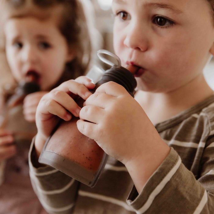 Two young children sip from bottles with straws, beginning their self-feeding journey. The child in the foreground wears a striped shirt and holds the drink close, while another enjoys a Haakaa Reusable 100% Food Grade Silicone Baby Food Pouch in the slightly blurred background.