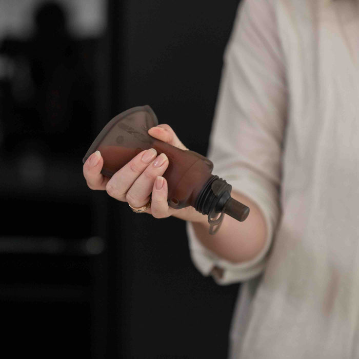 Someone holds a small, brown, translucent Haakaa squeeze bottle with a black spout against a dimly lit background, echoing the planet-friendly design of Reusable 100% Food Grade Silicone Baby Food Pouches.