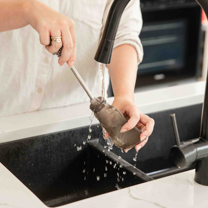 At a kitchen sink, someone uses a bottle brush to clean a baby bottle under running water. The brown liquid indicates deep cleaning. Haakaa Reusable 100% Food Grade Silicone Baby Food Pouches sit nearby, ready for their next eco-friendly use on the white countertop with an elegantly prominent black faucet.