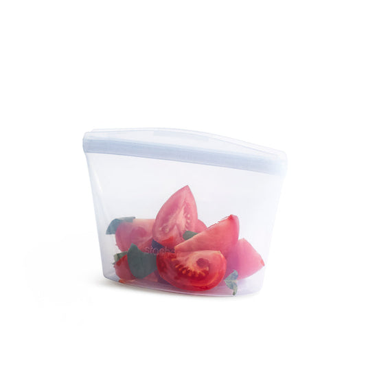 A semi-transparent, food-grade silicone bag holds sliced tomatoes and basil leaves, resembling a compact Stasher Reusable 2-Cup Bowl, set against a pristine white background.
