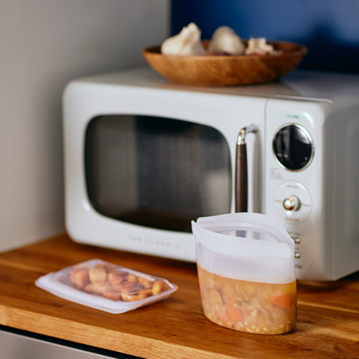 Stashers Reusable 2-Cup Bowls filled with meals are on a wooden countertop before a white microwave, offering a tidy alternative to traditional to-go containers. A plate with garlic bulbs is atop the microwave, complemented by the calming blue wall in the background.