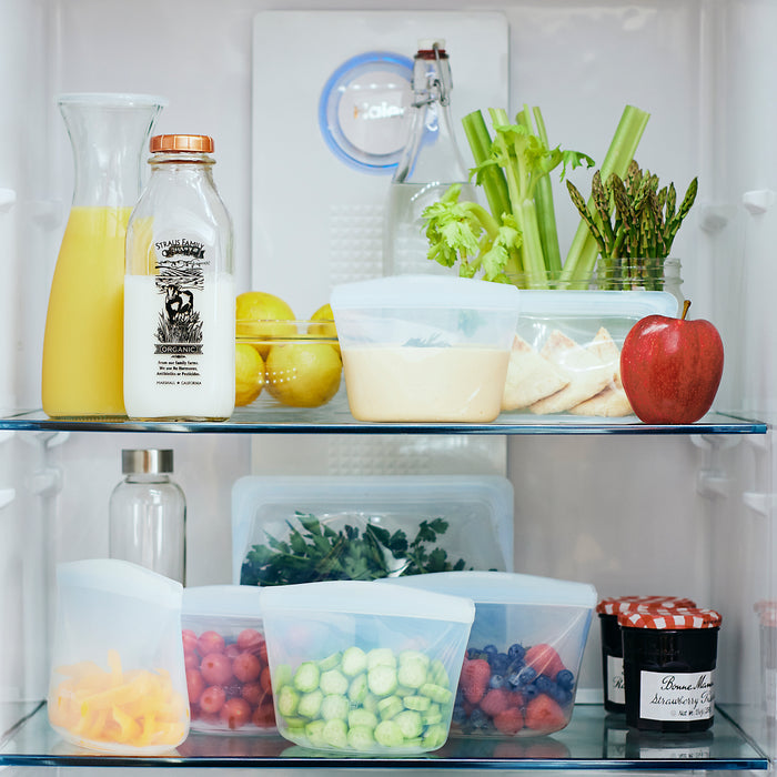 An open fridge reveals orange juice, milk, celery, asparagus, lemons, an apple, hummus, and pita on the top shelf. The bottom shelf has leafy greens in containers, grapes, pickles, a jar of jam, and carrots stored in a Stasher Reusable 2-Cup Bowl made from food-grade silicone.