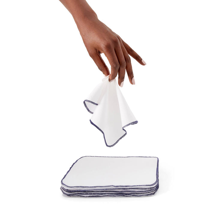 A hand selects a white cloth napkin with blue edges from a pile of Esembly Babys Reusable Baby and All Purpose Wipes, made from certified unbleached 100% organic cotton, set against a plain white background.