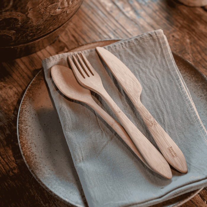 The Bambu Reusable Bamboo Spoon, Knife & Fork Set sits on a folded grey napkin placed atop a ceramic plate on a rustic wooden table.