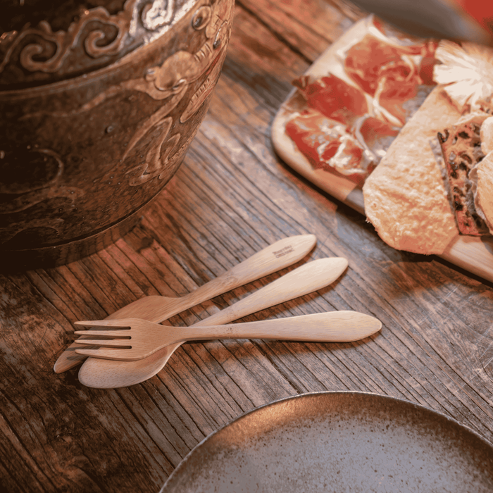 A Bambu Reusable Bamboo Spoon, Knife & Fork Set is arranged on a rustic wooden table beside a ceramic bowl and a platter of cured meats and crackers, with a ceramic plate partially visible in the foreground.