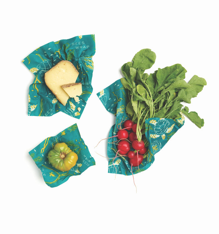 The Bees Wrap Assorted Wraps, sustainable food storage solutions, hold a wedge of cheese, a green tomato, and radishes. They feature a teal color with abstract yellow designs beautifully laid on a white background.