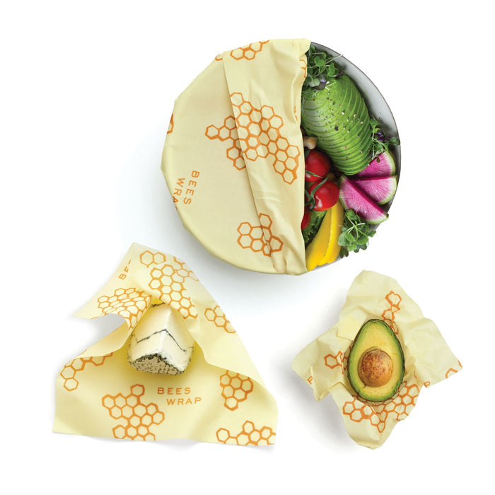 Three food items—half an avocado, mixed vegetables in a round container, and a wedge of cheese—are wrapped in Bees Wrap Assorted Wraps featuring honeycomb patterns and Bees Wrap branding.