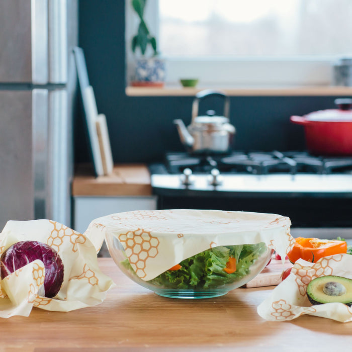A kitchen counter displays bowls and produce covered with Bees Wrap Assorted Wraps for eco-friendly food storage; a stove, kettle, and red pot appear in the background.