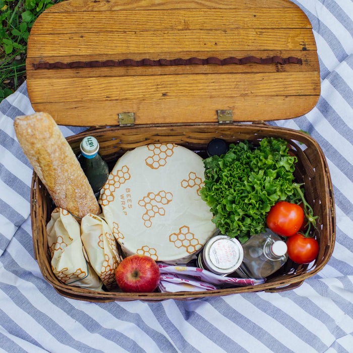 An open wooden picnic basket on a striped cloth, filled with food and drinks, features Bees Wrap Assorted Wraps by Bees Wrap for eco-friendly storage of items like a baguette, lettuce, tomatoes, apple, and jars.