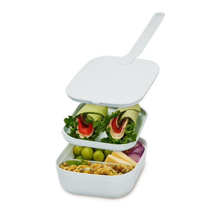 The W&P Reusable Bento Box is a portable, two-tier lunch container with compartments for wrap halves with lettuce and tomato on top, and grapes, cheese slices, onion slices, plus grain salad on the bottom.