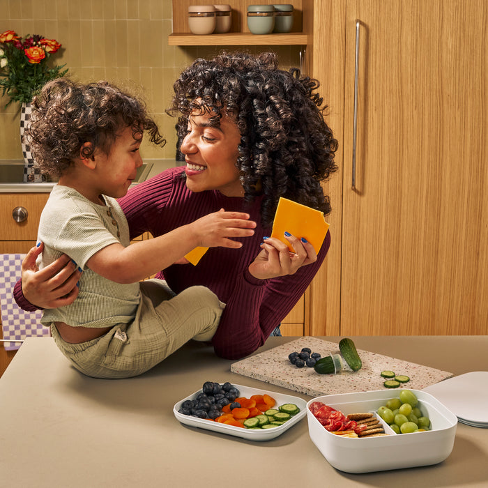 A woman with curly hair holds cheese, smiling at a toddler on the kitchen counter. The child grins, reaching for snacks in a W&P Reusable Bento Box filled with grapes, cucumbers, and crackers. The kitchen is warmly lit with wooden cabinets.