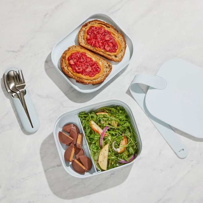 A Reusable Bento Box by W&P in light blue rests on a marble surface, featuring multiple compartments: one with two slices of bread and tomato spread, another with salad, avocado, orange slices, and peanut butter cups. A fork and spoon are neatly tucked in a blue holder.