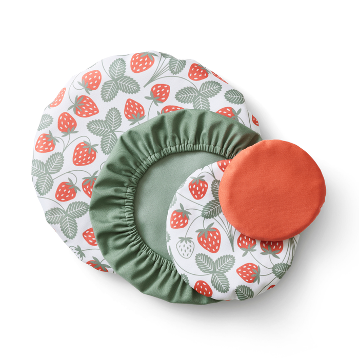 Four Esembly Baby Bowl Caps, crafted from recycled plastic, are displayed in a circular pattern. Three have a strawberry and green leaf design, while one is solid orange. These machine-washable covers blend style with eco-friendliness.