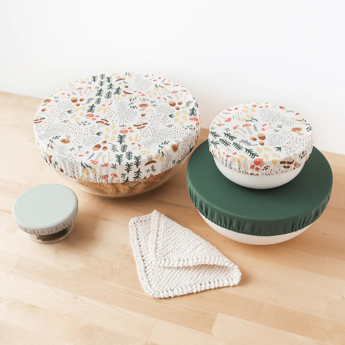 Three bowls of assorted sizes are topped with Esembly Baby Bowl Caps, made from recycled plastic, featuring autumn leaf patterns; two have floral designs and one is solid green. A folded knit cloth nearby on the wooden surface highlights an eco-friendly choice.