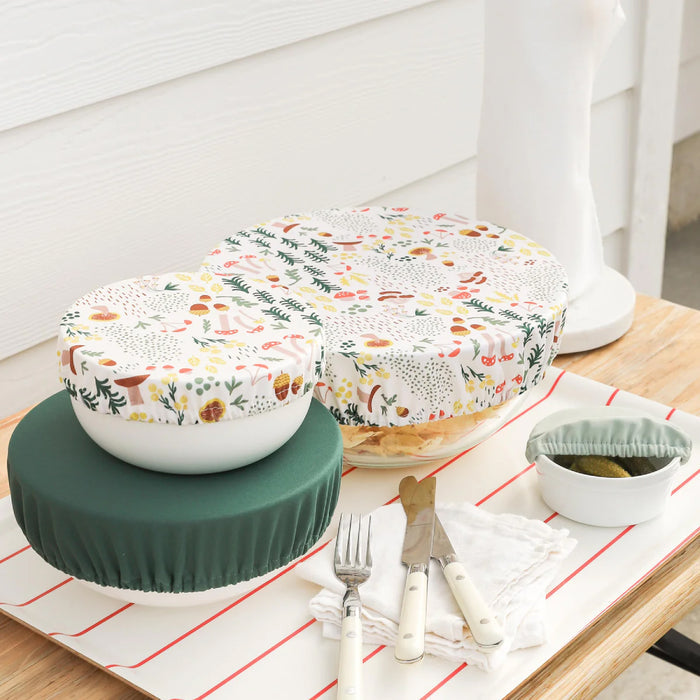 On a wooden table with a white cloth, two forks and a knife accompany bowls featuring machine-washable floral and green fabric covers. A small dish with an Esembly Baby Bowl Cap sits nearby, all against a pristine white wall backdrop.