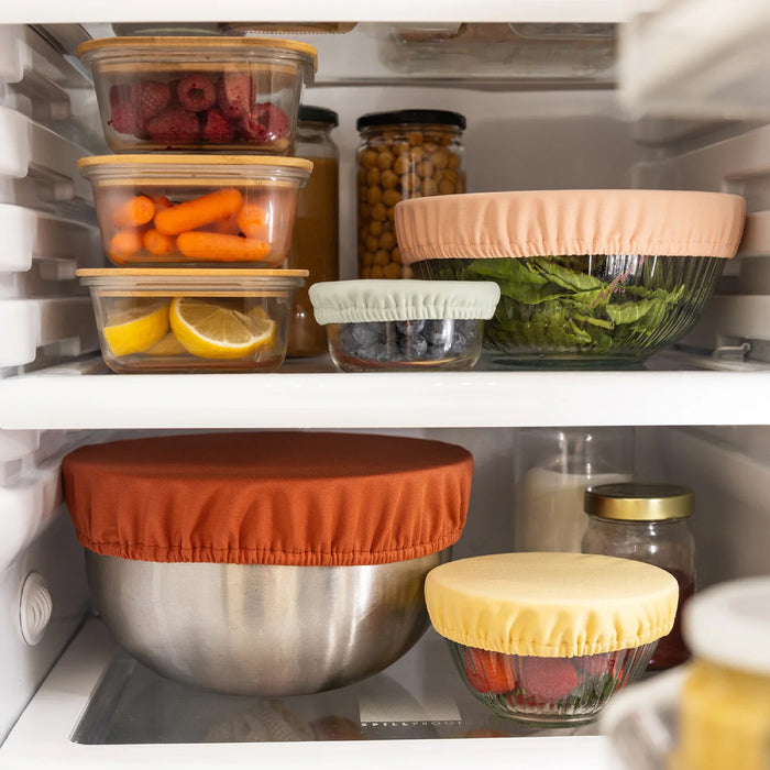 Refrigerator shelves house bowls with Esembly Babys Bowl Caps made from recycled plastic. They cover raspberries, carrots, chickpeas, greens, blueberries, lemon slices, and jars. The elastic fabric caps are machine-washable for added convenience.