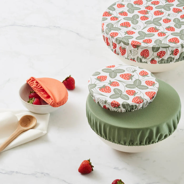 Three bowls feature Esembly Babys Bowl Caps: machine washable, sustainable covers with strawberry and green leaf designs, a solid green cover, and a small orange cover. A wooden spoon, napkin, and scattered strawberries are on the white marble surface.