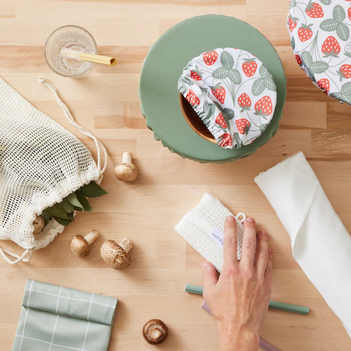 A table adorned with Esembly Baby Bowl Caps in strawberry patterns, a machine-washable cloth held by hand, and a leaf-themed mesh produce bag. Mushrooms rest near a strawed glass. A white cloth and checked fabric add to the eco-friendly display.