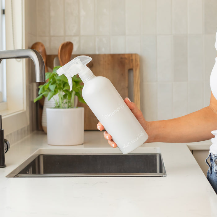 Someone holds a Cleanery Reusable Cleaning Bottle above a kitchen sink. In the background, a sugar cane cutting board sits next to a potted plant on the countertop, complemented by light tiles and a sleek faucet for an eco-friendly vibe.