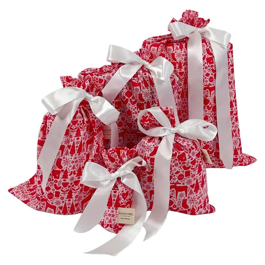 Five festive red and white patterned gift bags from the Waste Free Celebrations Reusable Gift Bags Set—crafted from 100% cotton—are displayed in a charming arrangement, each tied with a large, elegant white satin ribbon bow. A small Waste Free Celebrations tag is attached to the smallest bag at the front, showcasing these beautiful bags as an eco-friendly and sustainable alternative to traditional gift wrap for any special occasion.