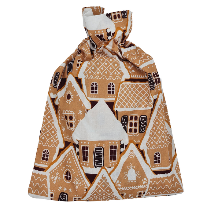 The Reusable Gift Bag from Waste Free Celebrations features a gingerbread house design with icing details on windows and doors. Made from 100% cotton, it serves as a festive and eco-friendly gift wrap option, secured at the top with a drawstring knot.