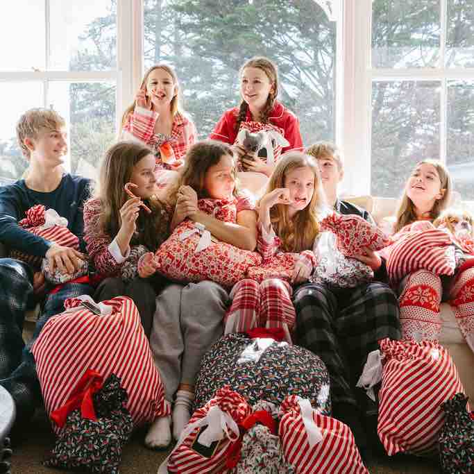 A group of kids in festive holiday pajamas sits on a couch, holding Waste Free Celebrations Reusable Gift Bags adorned with stripes and patterns. They smile, enjoying each others company as a large window with trees outside emphasizes their eco-friendly gift wrap choices.