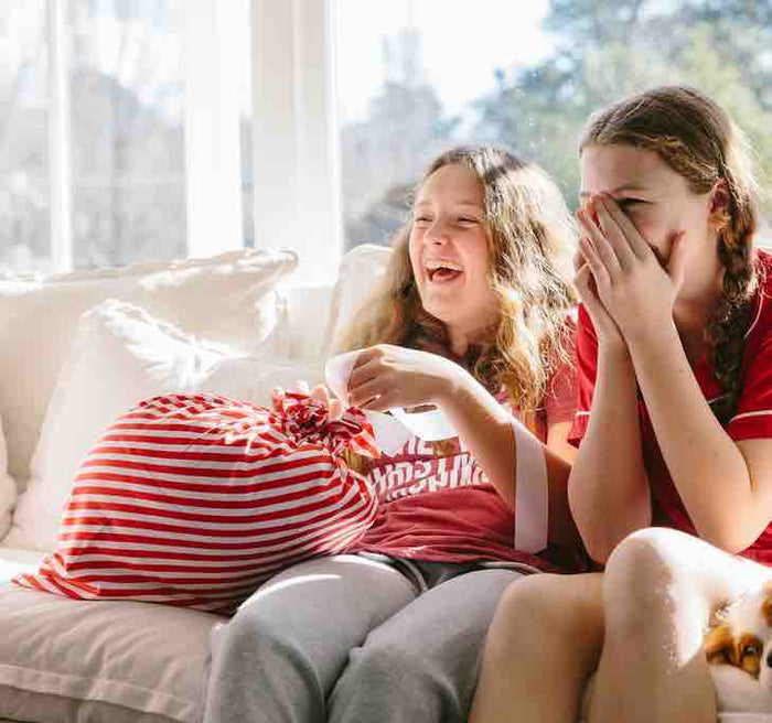 Two girls sit on a sofa, laughing in a brightly lit room. One holds a large red and white striped Reusable Gift Bag by Waste Free Celebrations, made of 100% cotton. The joyful atmosphere embodies the festive spirit of sustainable gift-giving.