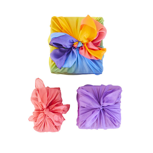 Three gift boxes wrapped in Sarahs Silks Reusable Gift Wrap Furoshiki Set with intricate knots: rainbow, pink, and purple patterns, each adorned with unique bows. Ideal for sustainable gift-giving against a white backdrop.
