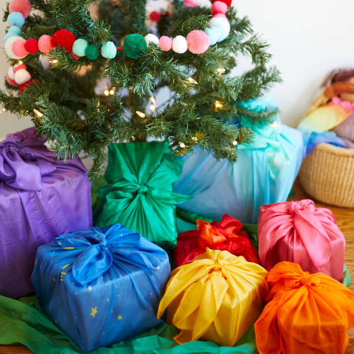 Vibrant Reusable Gift Wrap Furoshiki Sets by Sarahs Silks in purple, blue, green, red, yellow, and orange are wrapped around gifts under a Christmas tree adorned with a colorful pom-pom garland, highlighting sustainable gift-giving.