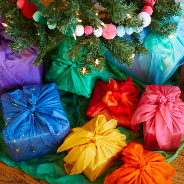 Under a decorated Christmas tree, gifts wrapped in Sarahs Silks Reusable Gift Wrap Furoshiki Set, featuring vibrant blue, yellow, red, green, and orange colors, add a sustainable touch amidst twinkling lights and ornaments.