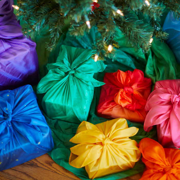 Vibrantly wrapped in the Reusable Gift Wrap Furoshiki Set by Sarahs Silks, colorful gifts in blue, green, red, yellow, orange, and purple rest under a Christmas tree, enhancing a festive atmosphere with sustainable gift-giving.