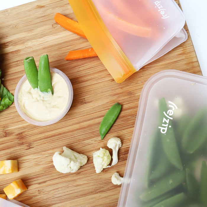 Zip’s Reusable Leakproof Bags Stand-Up Starter Kit holds carrot sticks, snap peas, and cheese cubes on a wooden cutting board, paired with a small cup of hummus and snap peas for a fresh snack setup.
