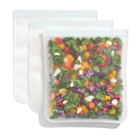 Three (re)zip Reusable Leakproof Gallon Bags are displayed: the front transparent bag holds chopped colorful vegetables, while two empty bags overlap behind it.