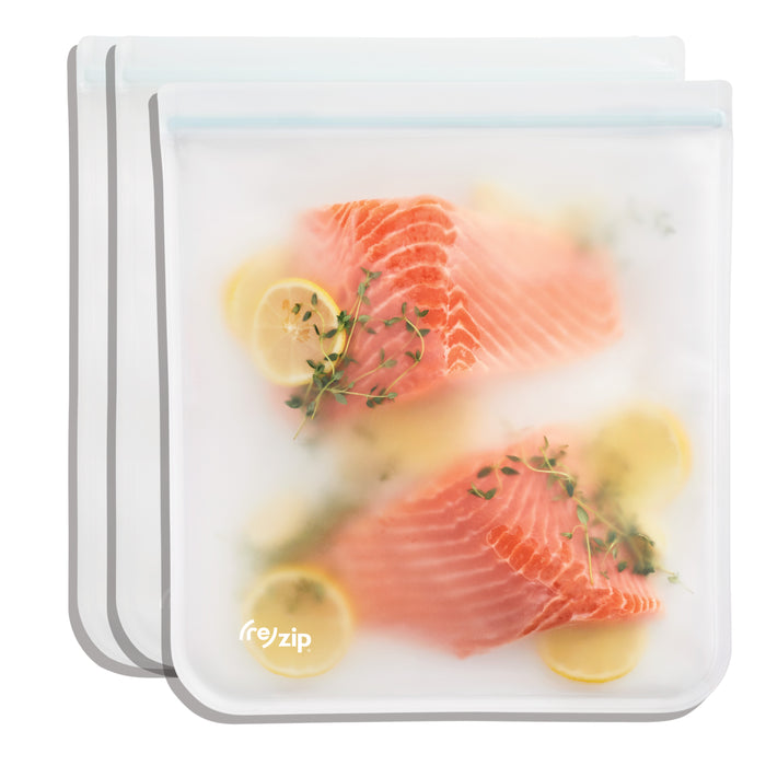 Three (re)zip Reusable Leakproof Gallon Bags, each filled with raw salmon slices, lemon slices, and herbs, are stacked behind one another and seen through a frosted surface.
