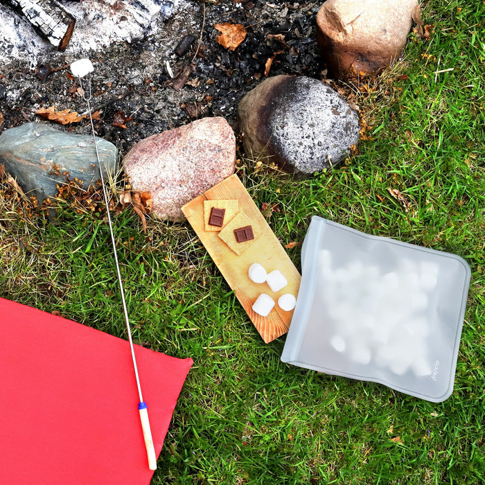 A campfire scene with a roasting stick over embers, chocolate and graham crackers on a wooden board, a (re)zip Reusable Leakproof Gallon Bag of marshmallows, and a red mat placed on green grass.