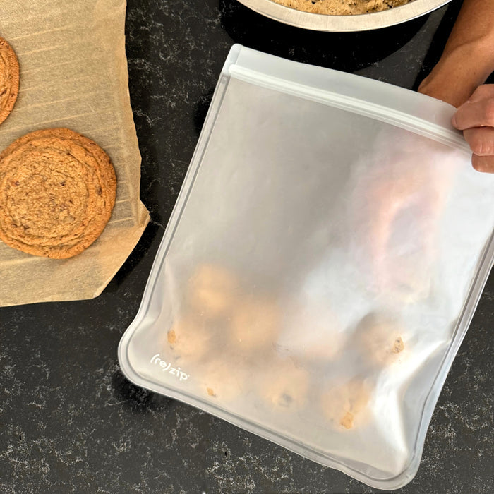 A hand holds a (re)zip Reusable Leakproof Gallon Bag with cookie dough balls inside, next to a tray of baked cookies and a baking sheet on a dark countertop.