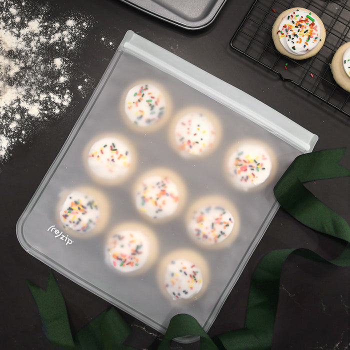 A (re)zip Reusable Leakproof Gallon Bag, freezer-safe, holds frosted sugar cookies with rainbow sprinkles on a flour-dusted dark countertop. Nearby are a cooling rack, baking tray, and a green ribbon.
