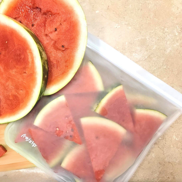 Slices of watermelon are on a light surface—some stacked on a cutting board, others kept fresh in (re)zip Reusable Leakproof Gallon Bags made from food-safe PEVA.