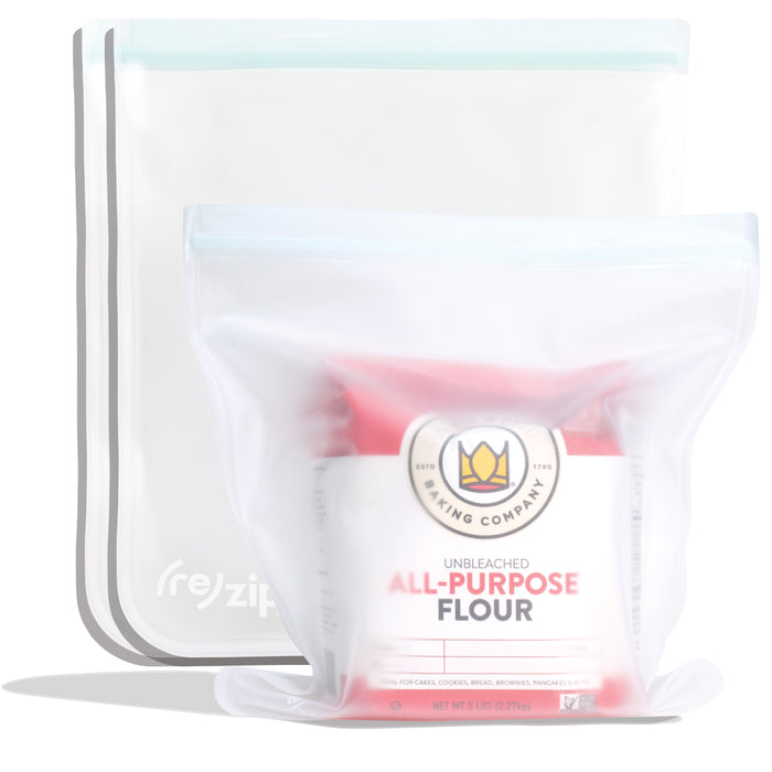 Two upright (re)zip Reusable Leakproof Gallon Bags, featuring transparent design and light blue seals, securely store a red and white bag of unbleached all-purpose flour. Perfect for freezer-safe food storage.
