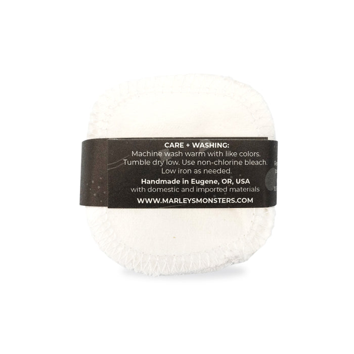 A white, fabric reusable cloth by Marleys Monsters with visible stitching and a black label featuring care instructions. Handmade in Eugene, OR, USA, from domestic and imported materials, these Reusable Organic Cotton Facial Rounds are perfect for sustainable facial care and gentle on the skin.
