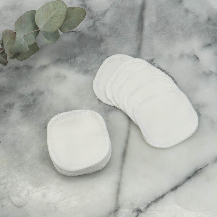 Marleys Monsters Reusable Organic Cotton Facial Rounds are stacked on a marble surface, with another set fanned out to the right. A eucalyptus sprig on the left emphasizes their connection to sustainable facial care.
