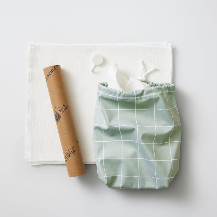 A green drawstring bag with a white grid pattern sits beside a rolled brown paper tube featuring a logo. An Esembly Baby Reusable Paper Towels Set and a small round object rest on the light surface, adding an environmentally conscious touch to the scene.