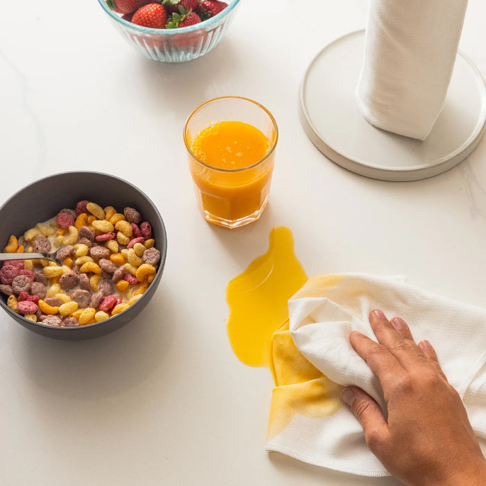 A persons hand uses Esembly Baby Reusable Paper Towels to clean spilled orange juice on a white table. Nearby are a bowl of colorful cereal with milk, a glass of orange juice, an eco-friendly paper towel holder, and a bowl of strawberries.