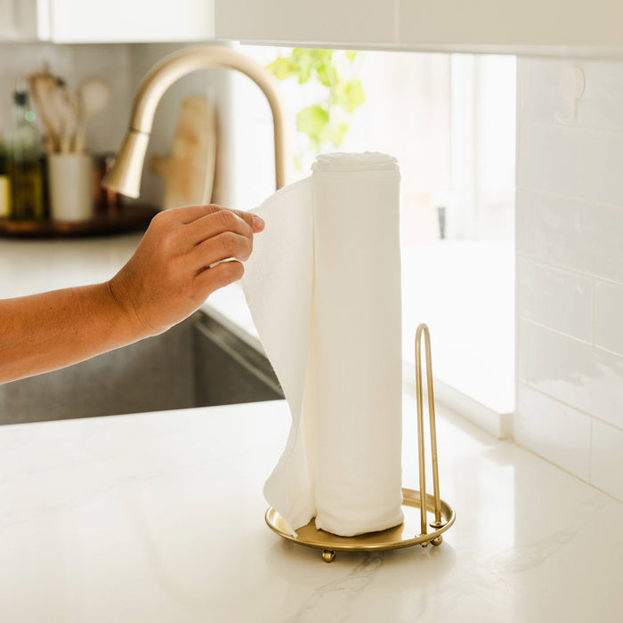 A hand pulls an Esembly Baby 100% Organic, Unbleached Reusable Paper Towel from a gold holder on a white kitchen countertop near a sink. The set includes 12 towels, 1 roll, 1 toss bag, and 1 hook.