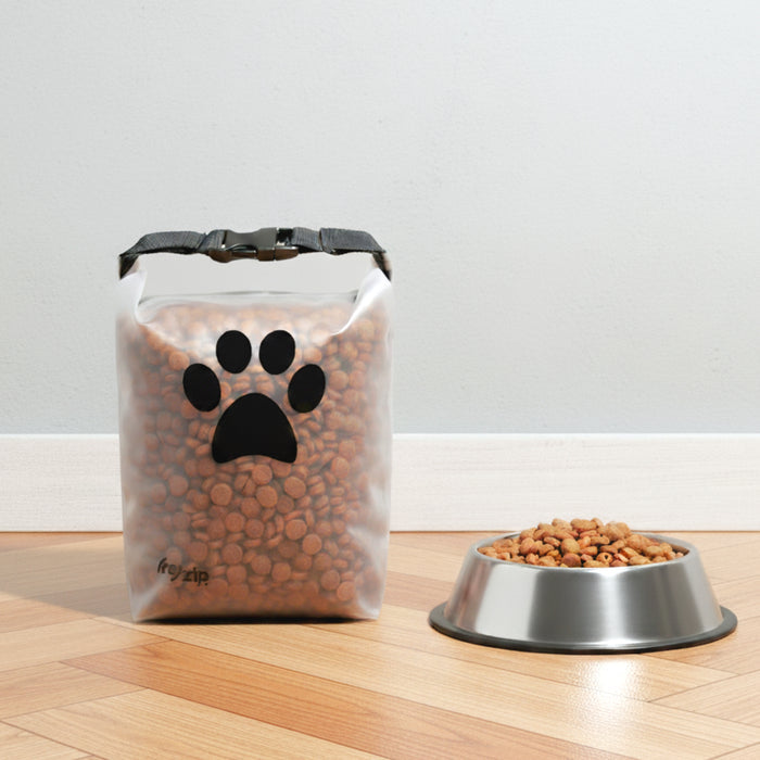 A (re)zip Reusable Pet Food Storage Bag - Medium, 14 Cup, with a black paw print design, sits on a wood floor filled with kibble beside a metal bowl of more kibble against a light gray wall.
