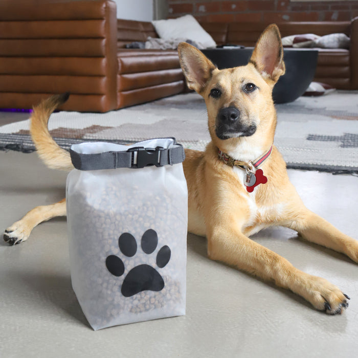 A tan dog with upright ears lies indoors near a (re)zip Reusable Pet Food Storage Bag - Medium, 14 Cup, featuring a black paw print—a perfect companion for storing kibble at home or on your next adventure.