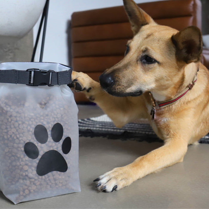 A tan dog lies on the floor, touching a (re)zip Reusable Pet Food Storage Bag - Medium (14 cup), filled with kibble and featuring a black paw print design. In the background are a brown couch and a patterned rug.